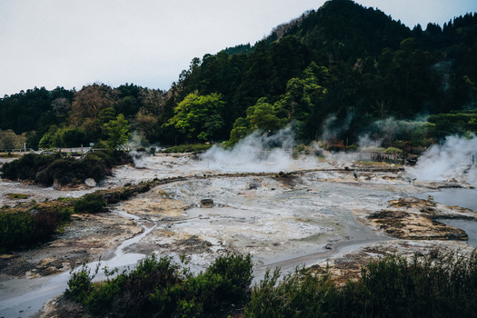 View of otherworldly steam rising from geothermal vents amidst the rugged mineral rich earth contrasting with the lush green hills Furnas Azores Portugal.