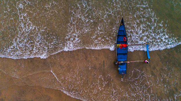 Aerial view of a traditional blue fishing boat on the sandy shore of Agonda Beach with waves washing over the sand and a fisherman standing nearby in Agonda Goa India.