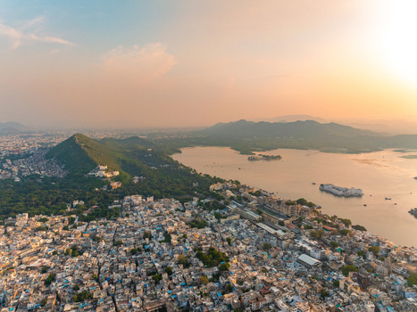 Aerial view of Udaipur featuring Lake Pichola the City Palace complex dense urban rooftops and surrounding Aravalli hills under a warm golden hour sky Udaipur Rajasthan India.