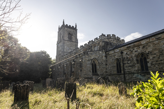View of the historic St. Oswald s Church with its stone tower rising above a weathered graveyard filled with ancient tombstones and tall grass in Durham England United Kingdom. View of the historic St. Oswald s Church with its stone tower rising above a weathered graveyard filled with ancient tombstones and tall grass in Durham England United Kingdom.