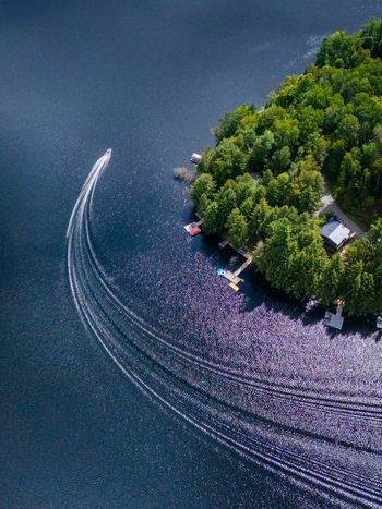 Aerial view of a motorboat leaving a long wake on the deep blue water of Baptiste Lake near a forested shoreline with a cottage in Hastings Highlands Ontario Canada.