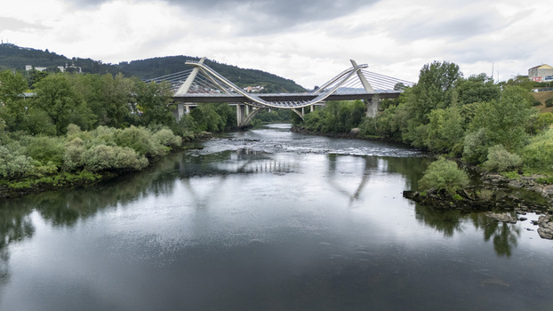 Aerial view of the modern Ponte do Milenio bridge spanning the Minho River with lush green riverbanks and rolling hills under a cloudy sky in Orense Ourense Spain. Aerial view of the modern Ponte do Milenio bridge spanning the Minho River with lush green riverbanks and rolling hills under a cloudy sky in Orense Ourense Spain.