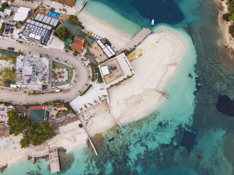 Aerial view of the turquoise waters and sandy beach of Ksamil with coastal buildings a wooden pier and small boats in Ksamil Vlore County Albania. Aerial view of the turquoise waters and sandy beach of Ksamil with coastal buildings a wooden pier and small boats in Ksamil Vlore County Albania.