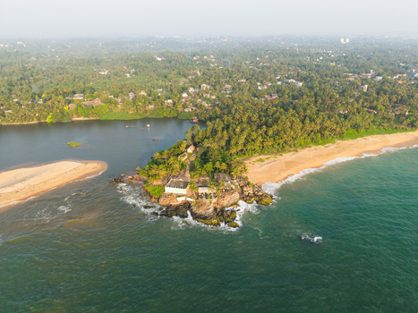 Aerial view of a rocky headland with a small white building golden sand beach and lush palm forests along the coastline in Galle Southern Province Sri Lanka.