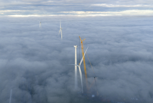 Aerial view of wind turbines and a yellow construction crane emerging from a thick layer of white clouds under an overcast sky.