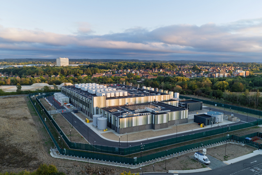 Aerial view of a modern data center facility with rooftop cooling units surrounded by a secure perimeter fence set within a suburban landscape in the United Kingdom. Aerial view of a modern data center facility with rooftop cooling units surrounded by a secure perimeter fence set within a suburban landscape in the United Kingdom.