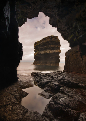 View of Dun Briste Sea Stack framed by the dark opening of a coastal cave with wet rocks and a puddle in the foreground in County Mayo Ireland. View of Dun Briste Sea Stack framed by the dark opening of a coastal cave with wet rocks and a puddle in the foreground in County Mayo Ireland.