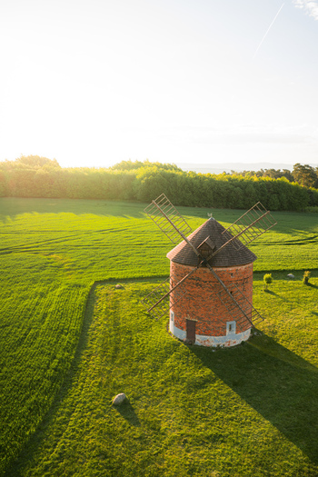 Aerial view of a historic brick windmill standing in a lush green field during golden hour with a dense forest in the background Chvalkovice Hradec Kralove Region Czechia.