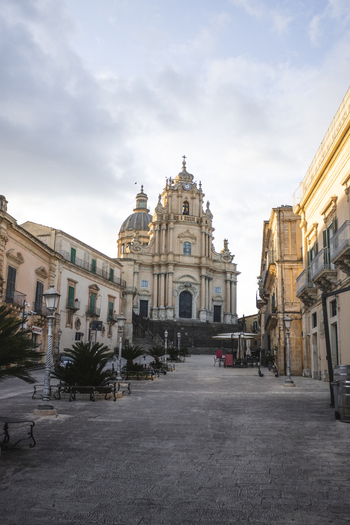 View of the Duomo di San Giorgio with its ornate Baroque facade overlooking a paved square and historic buildings under a soft sky in Ragusa Sicily Italy.