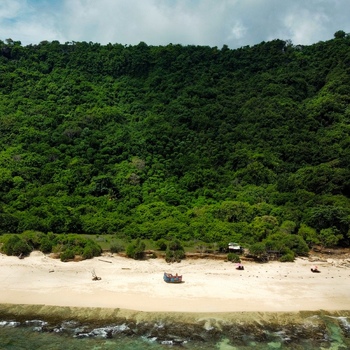 Aerial view of Pantai Nunggalan with a small boat on the white sand beach and dense green forest covering the cliffside in Kecamatan Kuta Selatan Bali Indonesia.