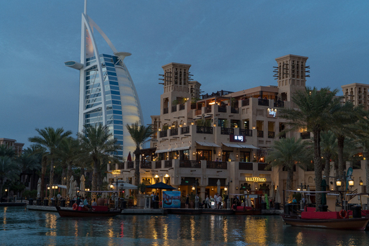 View of the Burj Al Arab luxury hotel and Madinat Jumeirah traditional buildings with palm trees and boats on the water at dusk in Dubai Dubai United Arab Emirates. View of the Burj Al Arab luxury hotel and Madinat Jumeirah traditional buildings with palm trees and boats on the water at dusk in Dubai Dubai United Arab Emirates.