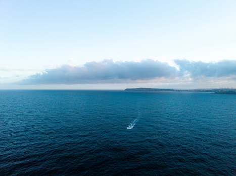 Aerial view of a small boat cruising on the deep blue ocean near the coast of Dee Why with a distant headland and soft clouds in the sky Dee Why New South Wales Australia. Aerial view of a small boat cruising on the deep blue ocean near the coast of Dee Why with a distant headland and soft clouds in the sky Dee Why New South Wales Australia.