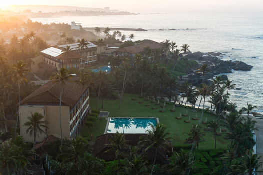 View of a luxury coastal resort with swimming pools and palm trees along the rocky shoreline at sunset in Galle Southern Province Sri Lanka.