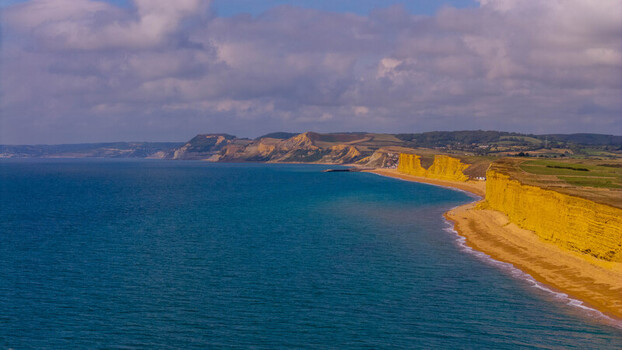 Aerial view of the dramatic golden sandstone cliffs meeting the blue waters of the English Channel under a cloudy sky in West Bay England United Kingdom. Aerial view of the dramatic golden sandstone cliffs meeting the blue waters of the English Channel under a cloudy sky in West Bay England United Kingdom.