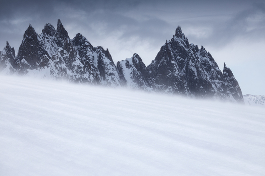 View of jagged dark mountain peaks rising above a vast wind swept snow field under an overcast sky in Aosta Valley Italy. View of jagged dark mountain peaks rising above a vast wind swept snow field under an overcast sky in Aosta Valley Italy.