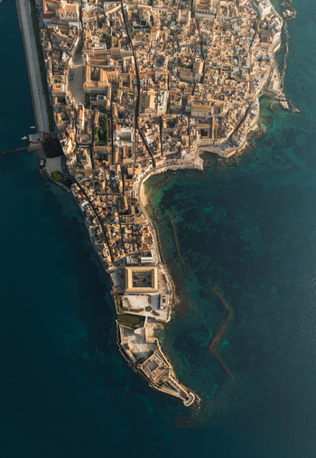 Aerial view of the historic Island of Ortigia featuring dense stone architecture and the Castello Maniace fortress surrounded by turquoise sea in Syracuse Sicily Italy.