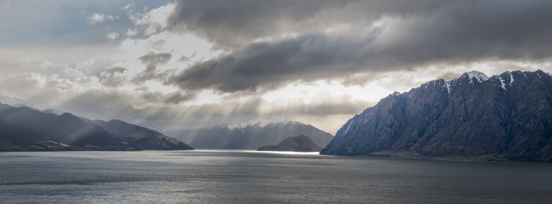 View of Lake Hawea surrounded by snow capped mountains under dramatic storm clouds with sunbeams breaking through in Otago Region New Zealand.