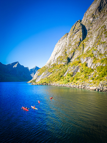 Aerial view of kayakers paddling through the deep blue waters of a fjord surrounded by steep rocky mountains and lush greenery in Hamnoy Nordland Norway.