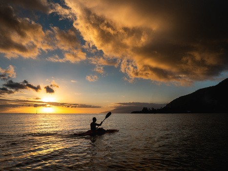 Aerial view of a person paddling a kayak on calm ocean waters during a golden sunset with dramatic clouds and the silhouette of Tahiti Moorea Maiao Windward Islands French Polynesia. Aerial view of a person paddling a kayak on calm ocean waters during a golden sunset with dramatic clouds and the silhouette of Tahiti Moorea Maiao Windward Islands French Polynesia.