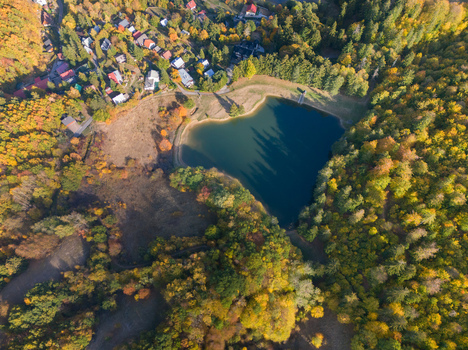 Aerial view of Pachomi tajch lake surrounded by dense autumn forest with colorful foliage and nearby houses in Stiavnicke Bane Banskobystricky kraj Slovakia.
