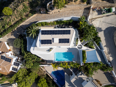 Aerial view of a modern villa featuring a swimming pool rooftop solar panels and lush palm trees in a residential neighborhood in Moraira Valencian Community Spain.