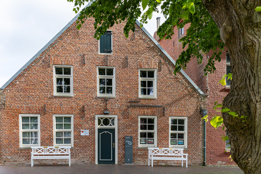 View of a traditional red brick building with white framed windows and benches under a leafy tree in Wittmund Lower Saxony Germany. View of a traditional red brick building with white framed windows and benches under a leafy tree in Wittmund Lower Saxony Germany.