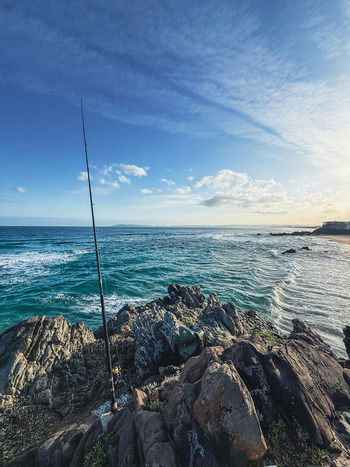 View of a fishing rod standing on rugged rocks overlooking crashing waves and the turquoise ocean under a blue sky in Plettenberg Bay Western Cape South Africa. View of a fishing rod standing on rugged rocks overlooking crashing waves and the turquoise ocean under a blue sky in Plettenberg Bay Western Cape South Africa.