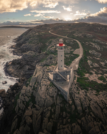 Aerial view of Faro de Punta Nariga lighthouse standing on rugged rocky cliffs with crashing waves and a winding coastal road under a cloudy sky in Malpica A Coruna Spain. Aerial view of Faro de Punta Nariga lighthouse standing on rugged rocky cliffs with crashing waves and a winding coastal road under a cloudy sky in Malpica A Coruna Spain.
