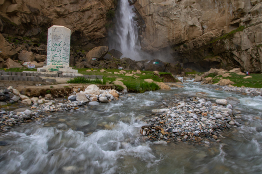 View of Khamosh Waterfall cascading down rugged cliffs into a rocky stream with a white monument in the foreground Skardu Gilgit Pakistan.