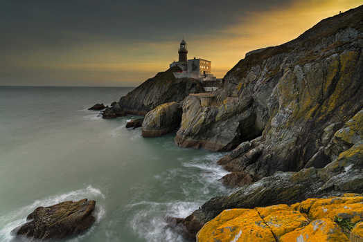 View of the Baily Lighthouse on the rugged cliffs of Howth with waves crashing against the rocks under a golden sunset sky in Ireland. View of the Baily Lighthouse on the rugged cliffs of Howth with waves crashing against the rocks under a golden sunset sky in Ireland.