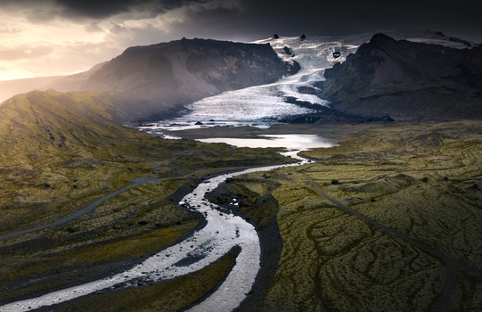 Aerial view of a massive glacier tongue flowing between rugged mountains into a winding river and glacial lake under a dramatic sky in Iceland.