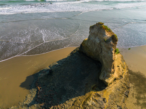Aerial view of a large rock formation on a sandy beach with gentle waves washing ashore under soft sunlight in Bali Bali Indonesia. Aerial view of a large rock formation on a sandy beach with gentle waves washing ashore under soft sunlight in Bali Bali Indonesia.