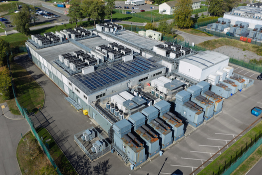 Aerial view of a data center facility with rooftop cooling units and solar panels surrounded by a fenced perimeter in the United Kingdom.