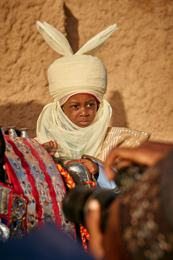 Zaria Nigeria 22 March 2026 View of a young person in a striking cream turban solemn against an earthy wall framed by vibrant regalia. Zaria Nigeria 22 March 2026 View of a young person in a striking cream turban solemn against an earthy wall framed by vibrant regalia.