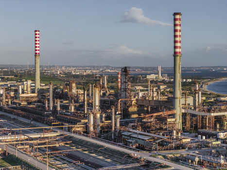 Aerial view of industrial complex with towering chimneys reaching towards the sky contrasting against the blue sea Syracuse Sicilia Italy.
