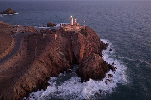 Aerial view of Lighthouse de Cabo de Gata perched on rocky cliffs above the sea at dusk with waves crashing against the shore and a winding coastal road in San Jose Andalucia Spain. Aerial view of Lighthouse de Cabo de Gata perched on rocky cliffs above the sea at dusk with waves crashing against the shore and a winding coastal road in San Jose Andalucia Spain.