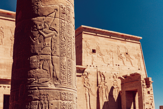 View of ancient Egyptian hieroglyphics and stone reliefs on a temple column with a pylon in the background under a clear blue sky at Philae Temple in Aswan Aswan Governorate Egypt. View of ancient Egyptian hieroglyphics and stone reliefs on a temple column with a pylon in the background under a clear blue sky at Philae Temple in Aswan Aswan Governorate Egypt.