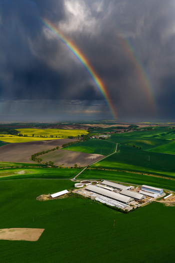 Aerial view of a farm complex surrounded by vibrant green and yellow agricultural fields under a dramatic stormy sky with a double rainbow in Nasedlovice South Moravian Region Czechia.