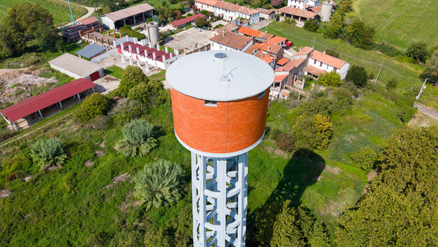 Aerial view of a water tower with a brick colored tank and concrete support structure surrounded by farm buildings lush green fields and trees in Mediglia Lombardy Italy. Aerial view of a water tower with a brick colored tank and concrete support structure surrounded by farm buildings lush green fields and trees in Mediglia Lombardy Italy.