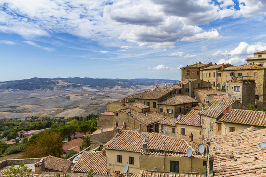 View of terracotta rooftops cascade down the hillside ancient stone buildings basking under a vast blue sky dotted with puffy white clouds Volterra Tuscany Italy. View of terracotta rooftops cascade down the hillside ancient stone buildings basking under a vast blue sky dotted with puffy white clouds Volterra Tuscany Italy.