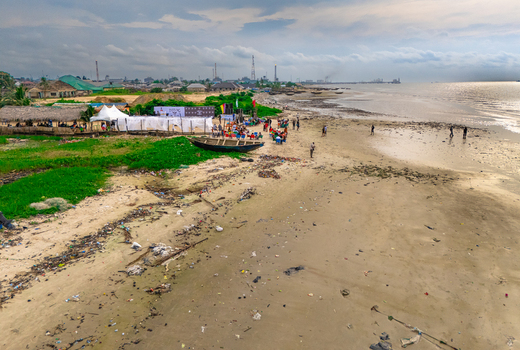 Aerial view of Bonny Island beach with a small wooden boat people gathered under white tents and industrial structures in the distance in Bonny Town Rivers Nigeria. Aerial view of Bonny Island beach with a small wooden boat people gathered under white tents and industrial structures in the distance in Bonny Town Rivers Nigeria.