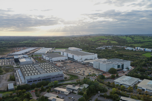 Aerial view of a large data center complex featuring industrial buildings parking lots and surrounding green fields under a cloudy sky in the United Kingdom.