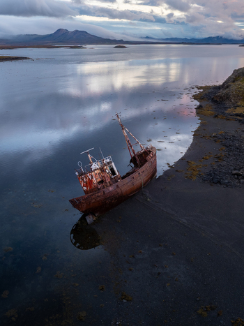 Aerial view of the stark rusty shipwreck resting on the black sand beach mirroring the moody sky with distant mountains Snæfellsnes Iceland. Aerial view of the stark rusty shipwreck resting on the black sand beach mirroring the moody sky with distant mountains Snæfellsnes Iceland.