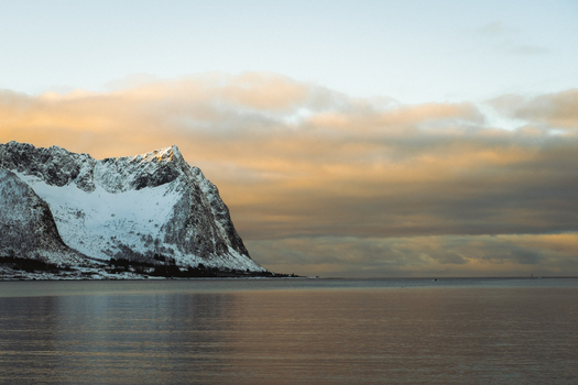View of snow capped mountain rises majestically from the tranquil waters under a sky streaked with warm hues of sunset Indregård Troms Norway. View of snow capped mountain rises majestically from the tranquil waters under a sky streaked with warm hues of sunset Indregård Troms Norway.