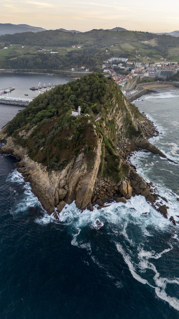 Aerial view of Getariako Itsasargia lighthouse perched on a rocky headland surrounded by crashing waves and lush greenery in Getaria Euskadi Spain. Aerial view of Getariako Itsasargia lighthouse perched on a rocky headland surrounded by crashing waves and lush greenery in Getaria Euskadi Spain.