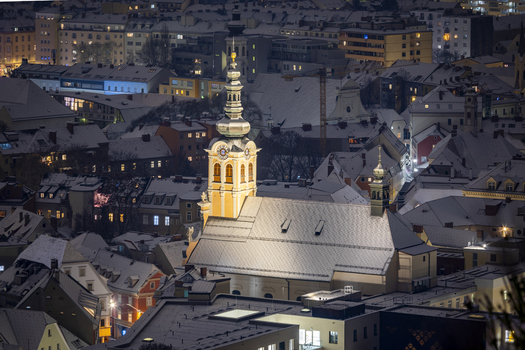 View of snow dusted rooftops and a brightly lit church tower pierce the twilight sky casting a serene glow over the winter landscape Graz Styria Austria. View of snow dusted rooftops and a brightly lit church tower pierce the twilight sky casting a serene glow over the winter landscape Graz Styria Austria.