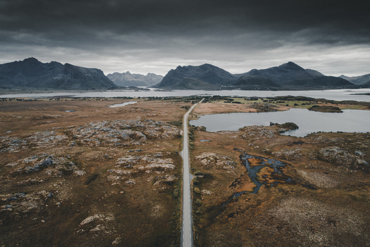 Aerial view of Gimsoy featuring a road cutting through rugged rocky terrain surrounded by lakes and dramatic mountains under a moody overcast sky in Vagan Municipality Nordland Norway. Aerial view of Gimsoy featuring a road cutting through rugged rocky terrain surrounded by lakes and dramatic mountains under a moody overcast sky in Vagan Municipality Nordland Norway.