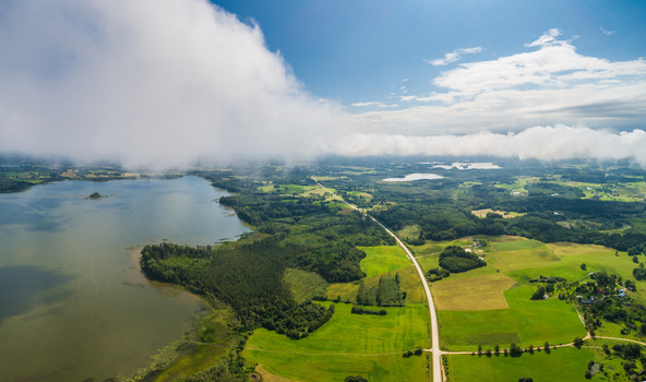 Aerial view of a serene landscape featuring a large lake lush green fields and a straight road cutting through the countryside under a sky filled with fluffy white clouds Latvia. Aerial view of a serene landscape featuring a large lake lush green fields and a straight road cutting through the countryside under a sky filled with fluffy white clouds Latvia.