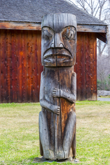 View of a traditional carved wooden totem pole standing in front of a rustic wooden building at Ksan Historical Village in Ksan British Columbia Canada. View of a traditional carved wooden totem pole standing in front of a rustic wooden building at Ksan Historical Village in Ksan British Columbia Canada.