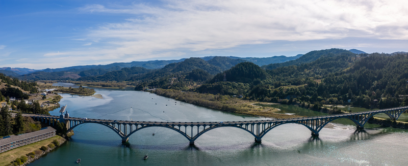 Aerial view of the Isaac Lee Patterson Bridge spanning the Rogue River with lush green hills calm water and a clear blue sky in Gold Beach Oregon United States. Aerial view of the Isaac Lee Patterson Bridge spanning the Rogue River with lush green hills calm water and a clear blue sky in Gold Beach Oregon United States.